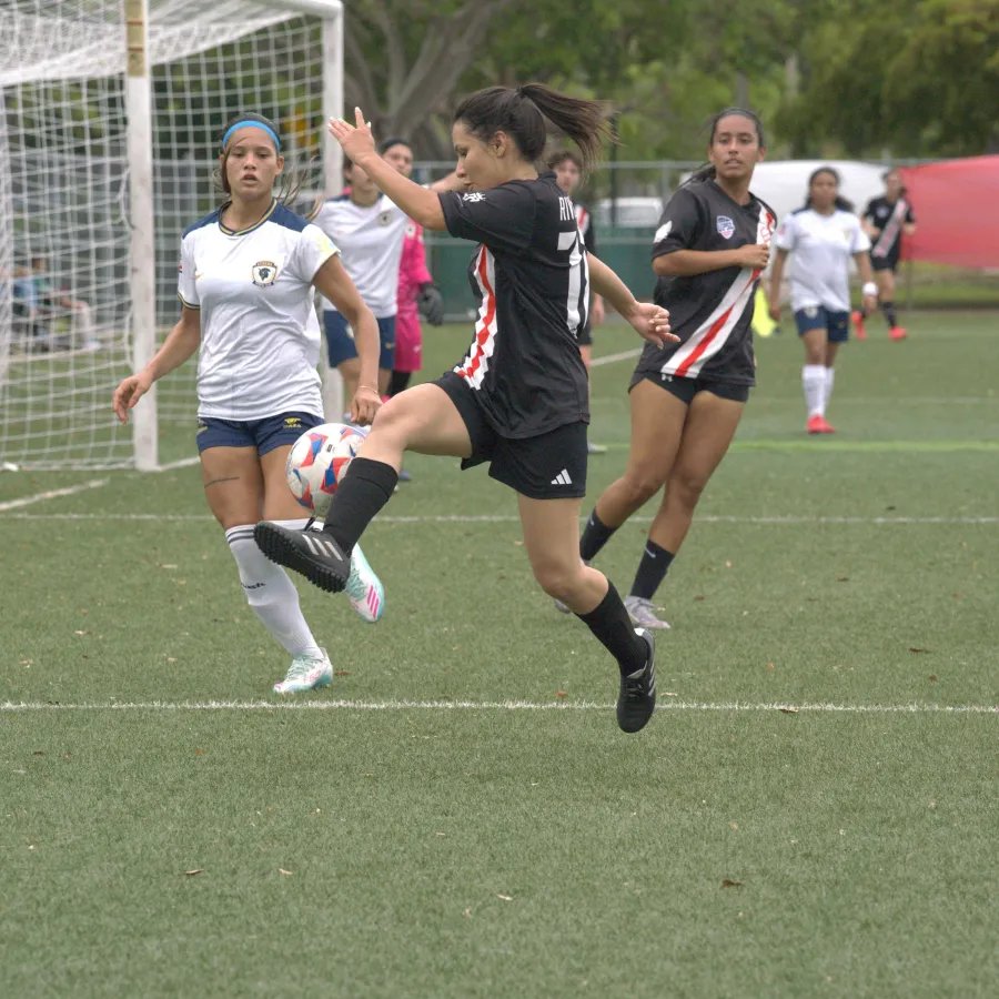 Female soccer player in black jersey kicking ball near goal while opponents in white defend on green field