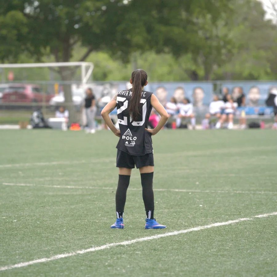 Female soccer player in black jersey standing on green field during match with goalpost and spectators in background
