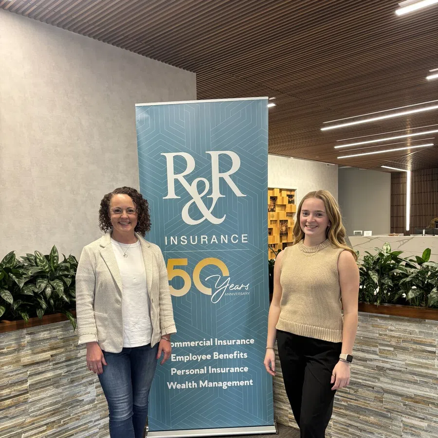 Two women standing beside an R&R Insurance 50 years anniversary banner in a modern office lobby.