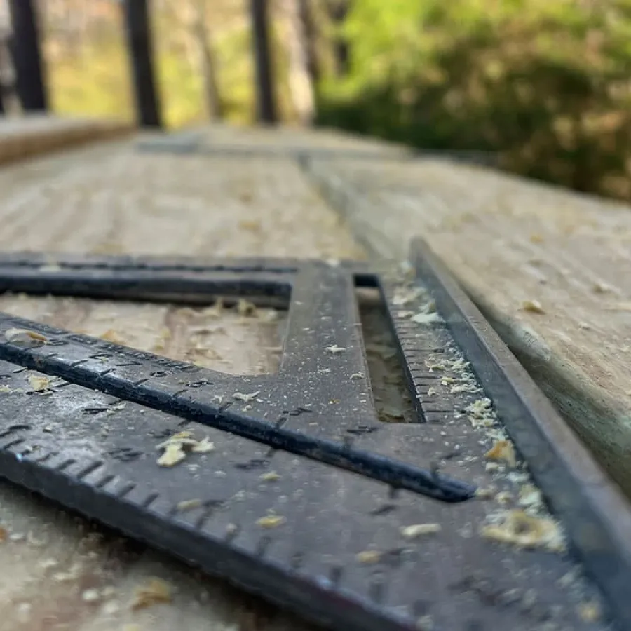 Close-up of a black carpenter's square on a wooden plank with sawdust in an outdoor wooded area.
