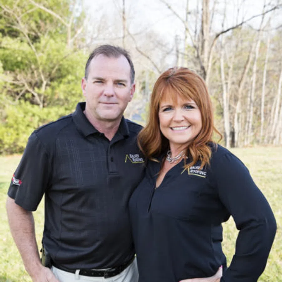 Man and woman standing outdoors in casual work attire smiling at the camera with greenery and trees in the background