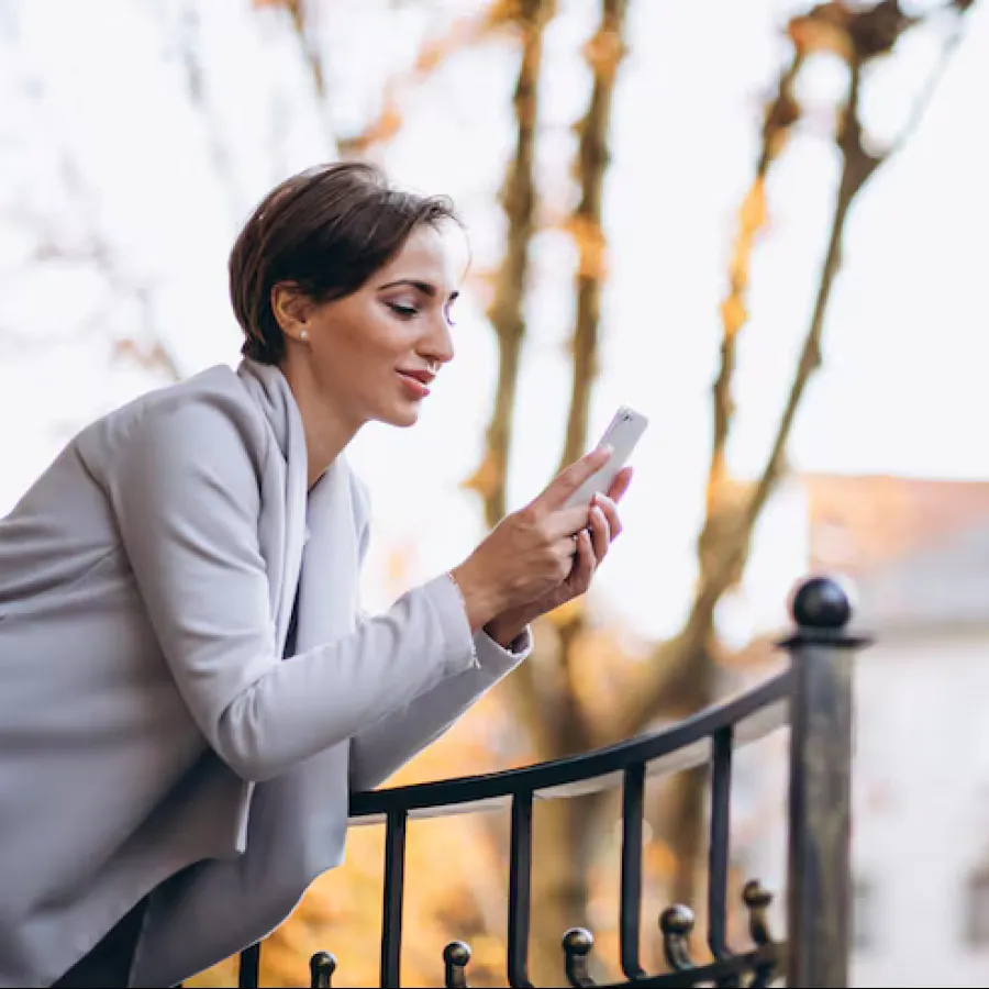 Woman in a gray coat leaning on a black iron fence using a smartphone outdoors during autumn.