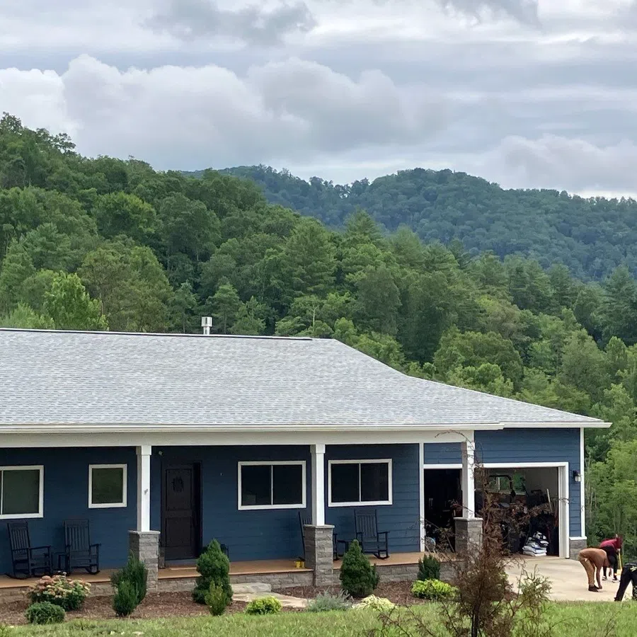 Blue single-story house with white trim and front porch surrounded by green trees and hills under cloudy sky