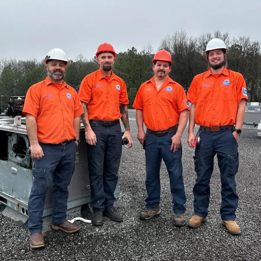 Four HVAC technicians in orange uniforms and hard hats standing on a rooftop beside HVAC equipment on a cloudy day