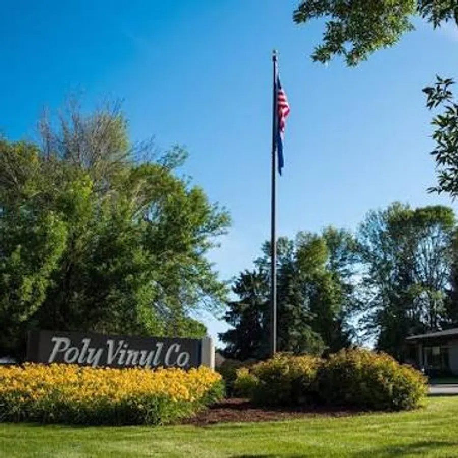 American flag flying on flagpole near Poly Vinyl Co sign surrounded by bushes and green trees on sunny day