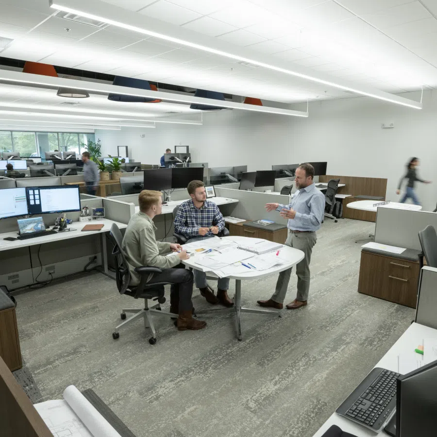 Modern open office space with employees collaborating at a round table surrounded by workstations and computers.