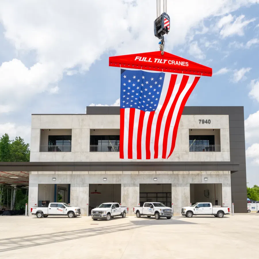American flag hanging from a Full Tilt Cranes hook above trucks in front of a modern industrial building.