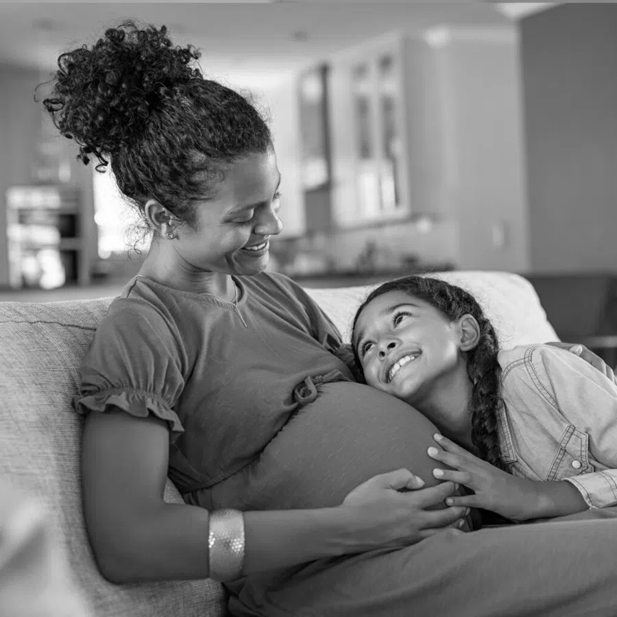 Smiling pregnant woman sitting on couch with daughter resting head on her belly in cozy home setting