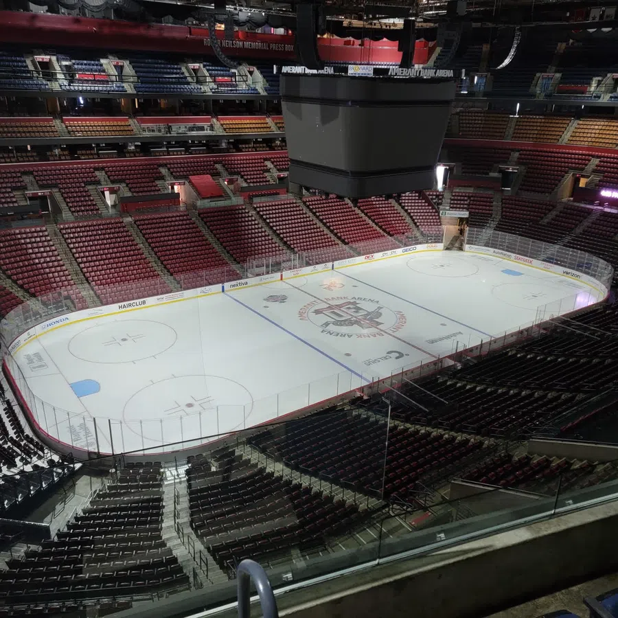 Empty indoor ice hockey arena with ice rink and surrounding red and black seats under bright lights.