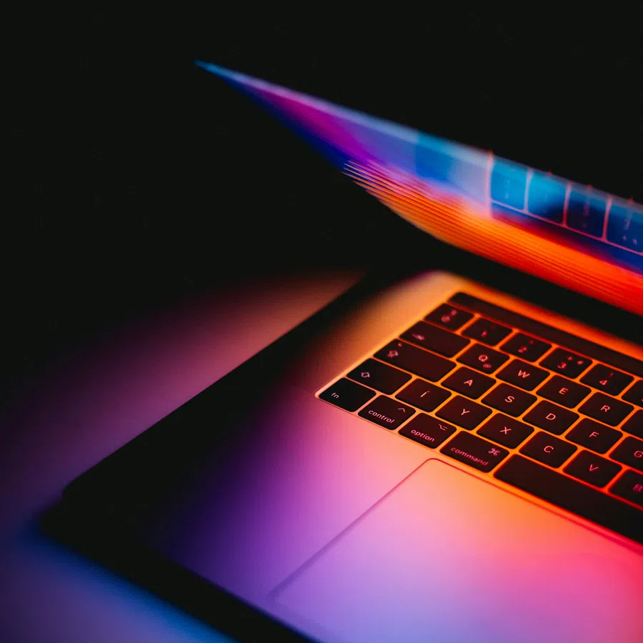 Partial view of a laptop keyboard illuminated by colorful neon lights in a dark environment.