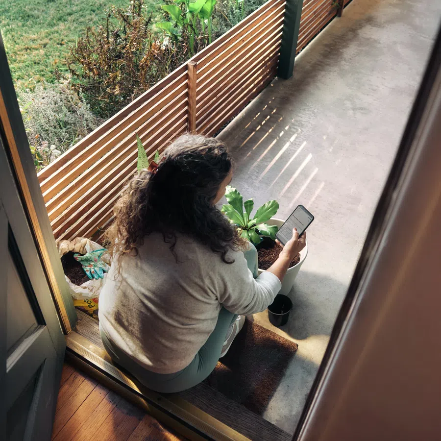 Woman sitting on doorstep checking phone with potted plants and garden tools around her in sunlight