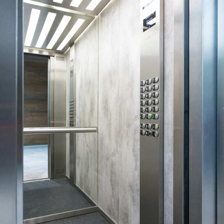 Interior of modern elevator with stainless steel panels, control panel, mirror, and strip ceiling lights.