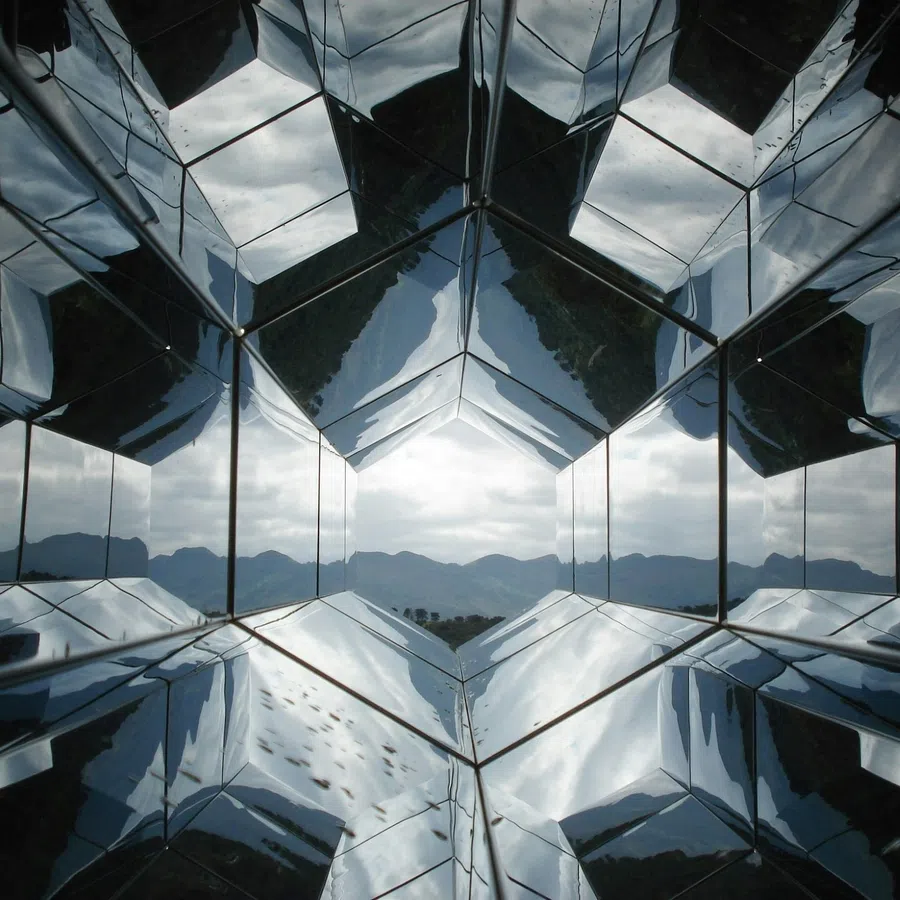 Geometric mirrored panels reflect a cloudy sky and mountains, creating a kaleidoscopic visual effect.