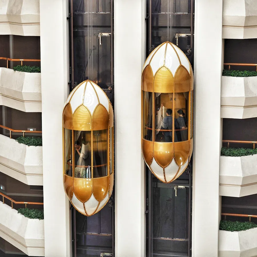 Two gold and white glass elevators ascend inside a multi-level modern atrium with terraces and plants.