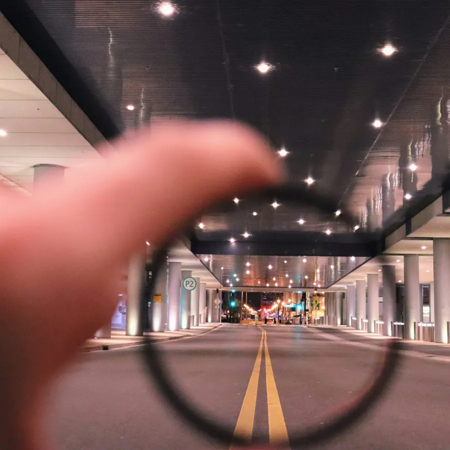 Hand holding a lens focusing on a city street under a modern, lit ceiling with pillars at night.
