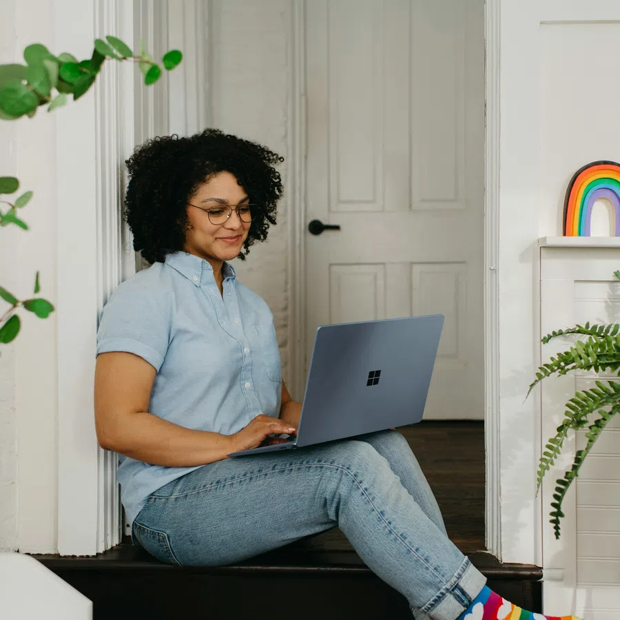 Woman with curly hair and glasses sitting on floor working on laptop in bright room with rainbow decor and green plant.