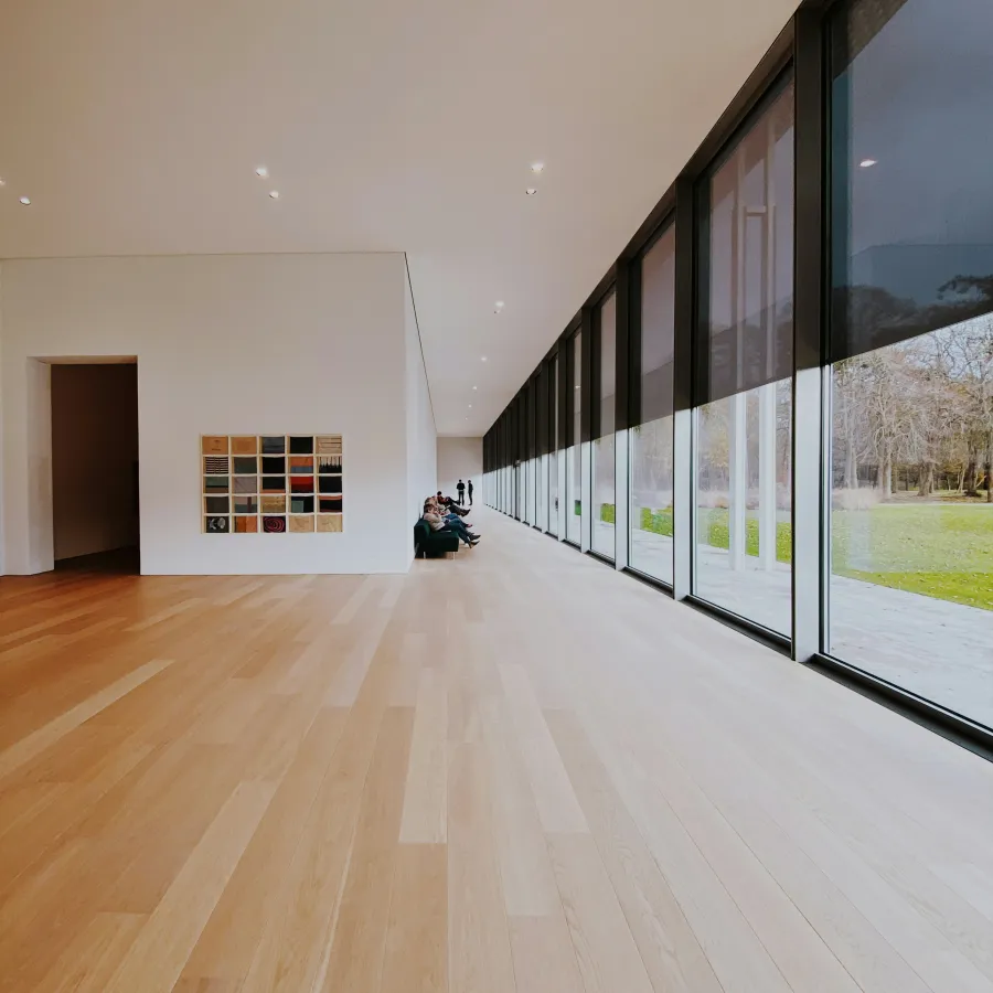 Minimalist gallery space with wooden floors, large windows with blinds, and people seated on a bench.