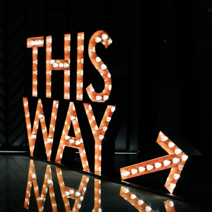 Illuminated marquee sign with words THIS WAY and arrow, reflecting on shiny black floor in dark room.