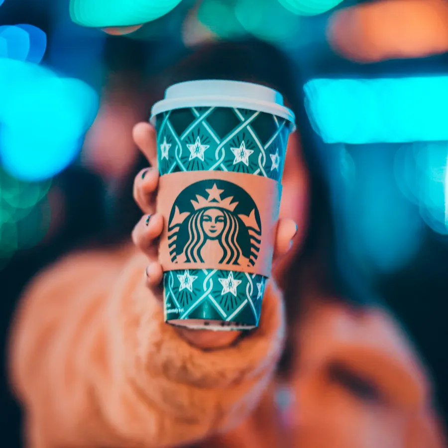 Hand holding a green Starbucks holiday cup with a blurred colorful bokeh background and person in orange coat.