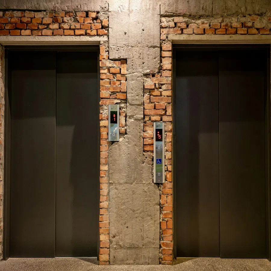 Two closed elevator doors framed by exposed brick and concrete walls, showing floor number displays.