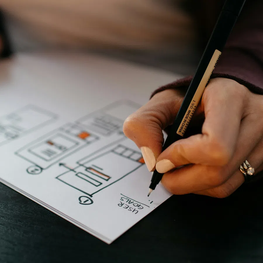 Close-up of a hand sketching a website user flow chart with a pen on white paper at a desk.