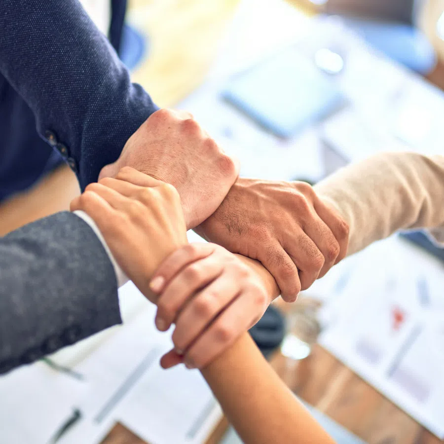 Four diverse business professionals joining hands in a teamwork gesture over an office desk with documents.