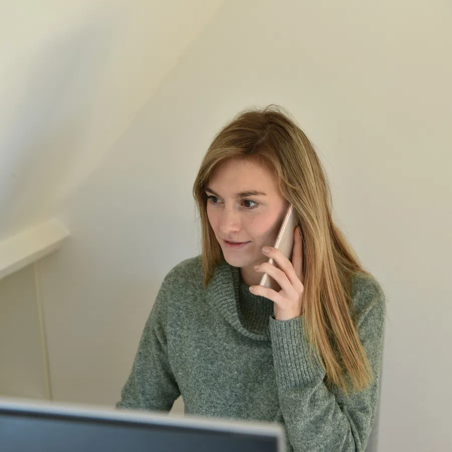 Woman in green sweater talking on phone while working at desk with computer in bright office space