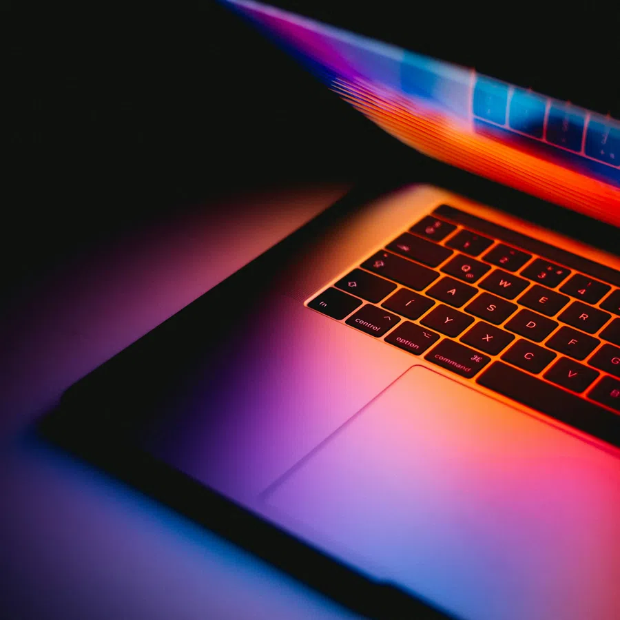 Partially open laptop keyboard illuminated by vibrant purple, orange, and blue lighting in a dark setting