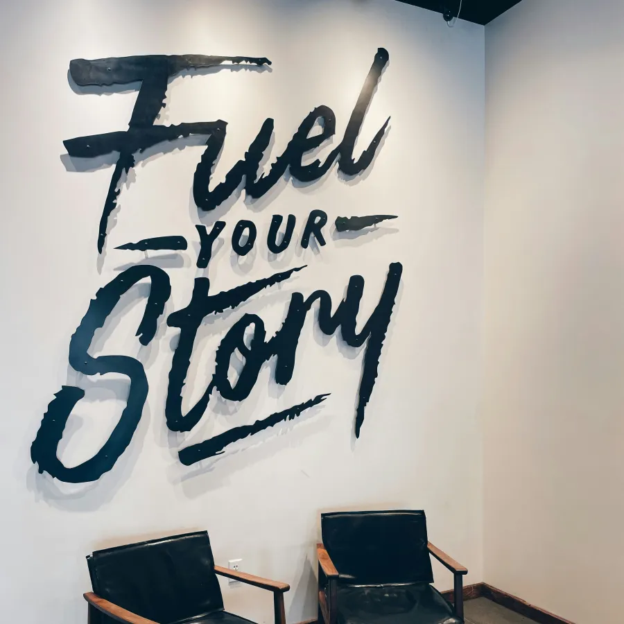 Cozy corner with two black leather chairs and wooden table under a wall sign saying Fuel Your Story in bold lettering.