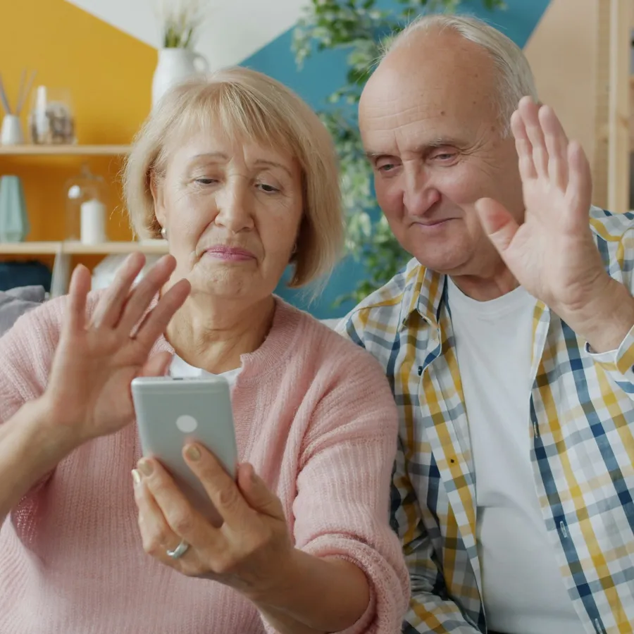 Senior couple video calling on smartphone waving hands in colorful living room.