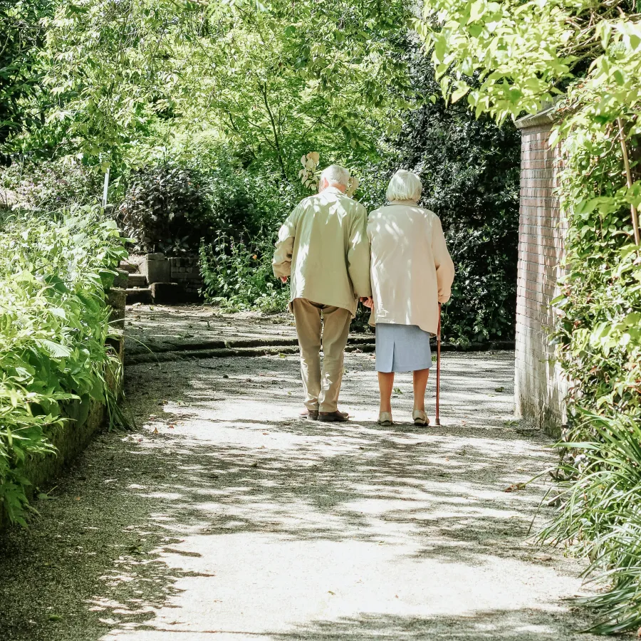 Elderly couple walking hand-in-hand on a shaded garden path surrounded by greenery.