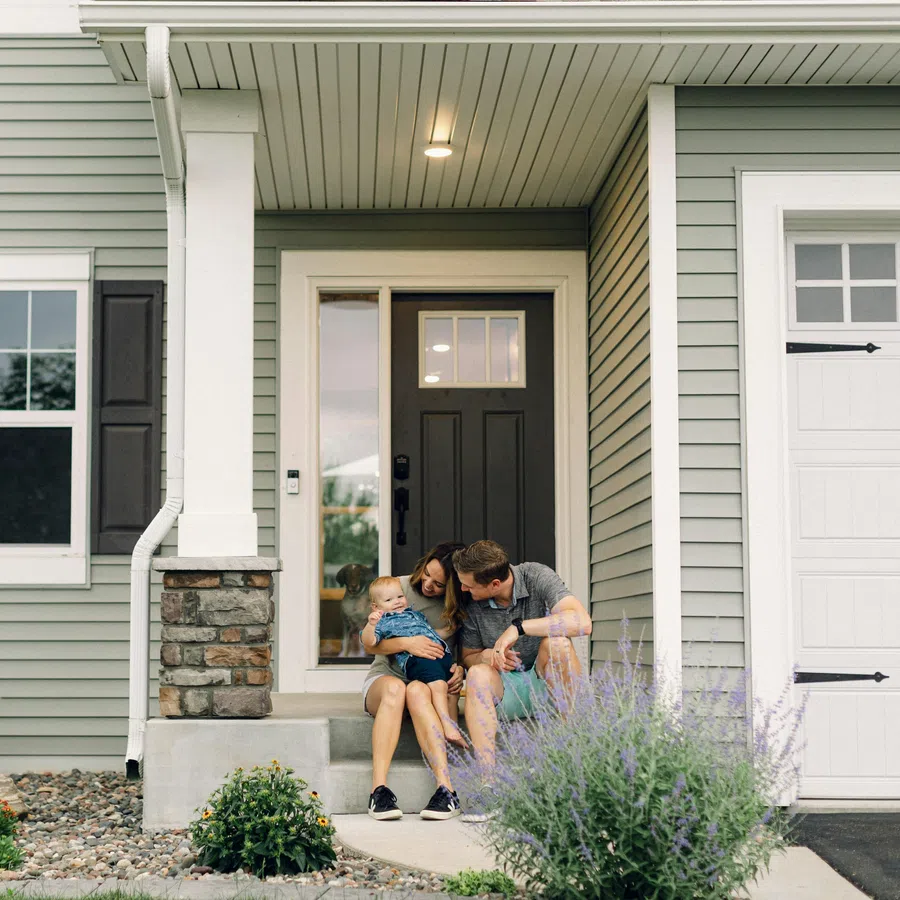 Family with toddler sitting on front porch steps of modern suburban house with stone accents and garden.