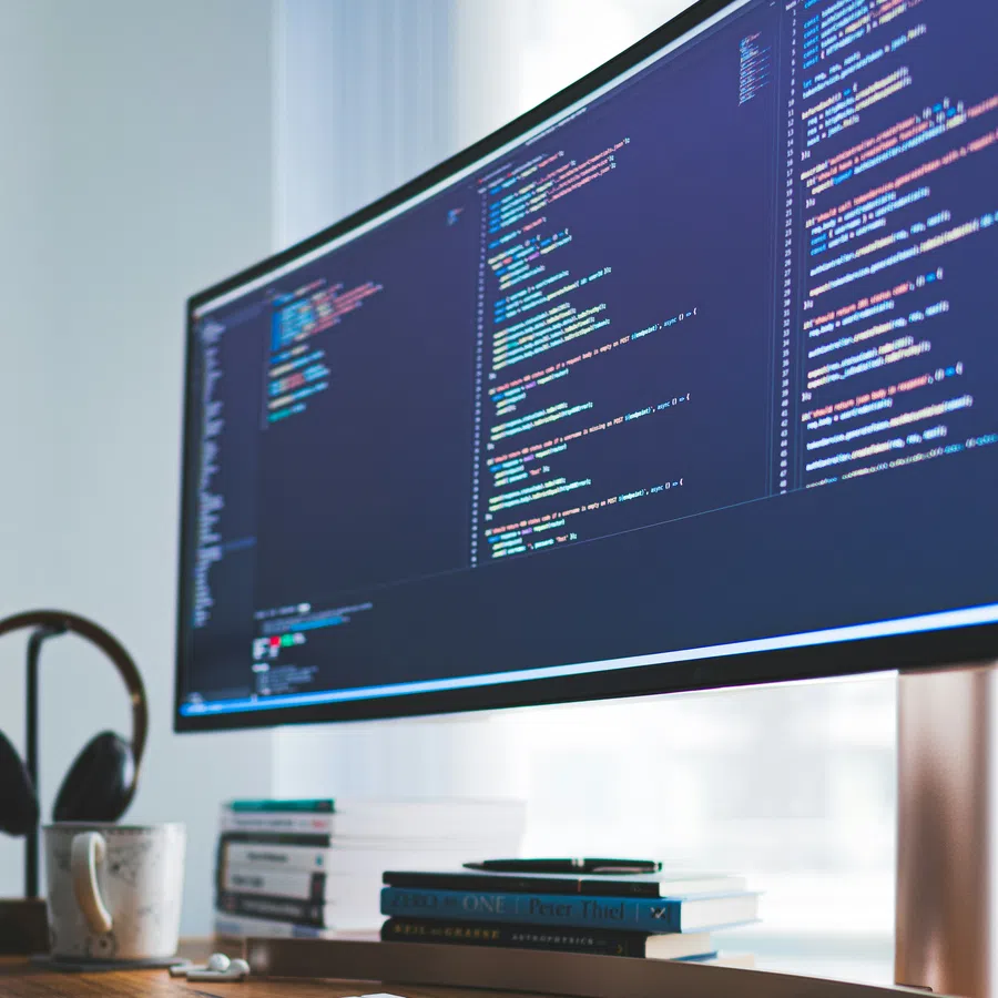 Wide computer monitor displaying colorful programming code on a desk with books, keyboard, and headphones.