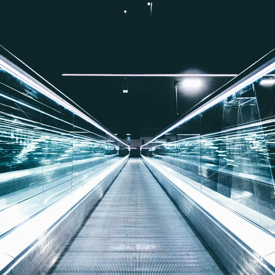 Futuristic illuminated walkway with glass railings and bright linear lights in a modern urban setting at night.