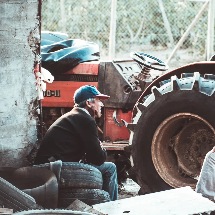 Two men wearing caps work beside a large red tractor in a rustic garage with old tires and tools.