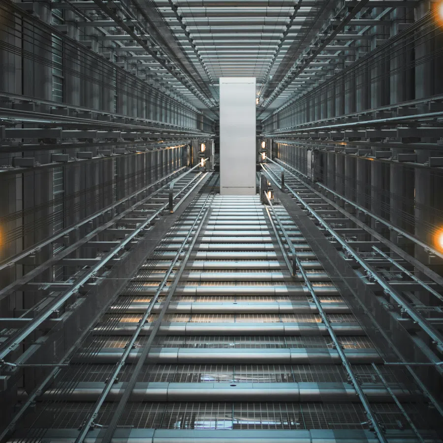 View looking up inside a modern glass elevator shaft with metal framework and warm lights illuminating the space