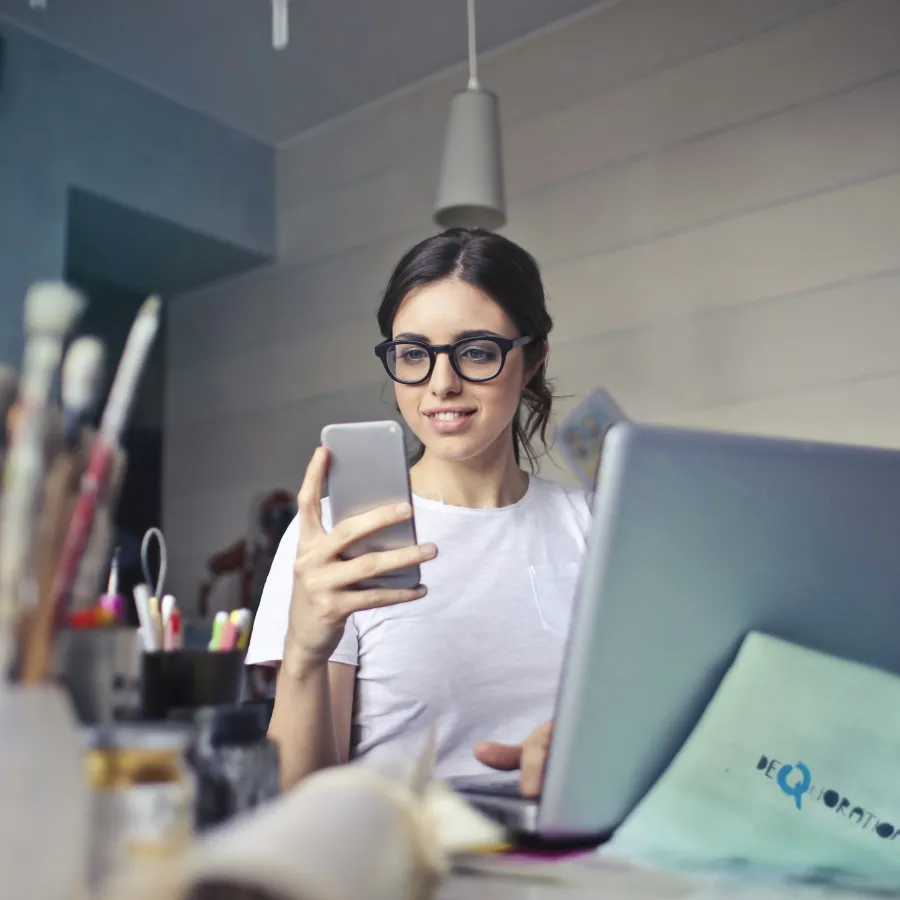 Young woman with glasses using smartphone and laptop in an art studio with brushes and supplies