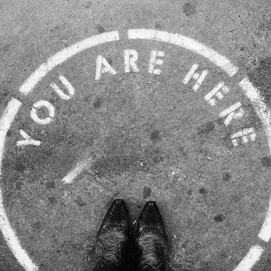Black and white photo showing feet inside a painted circle on pavement with words You Are Here.
