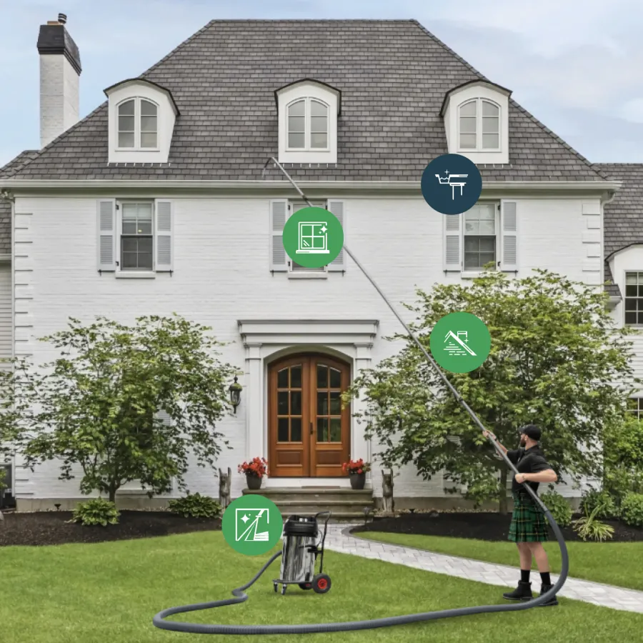 Man cleaning second-story windows of large white house using long pole vacuum tool on green lawn.