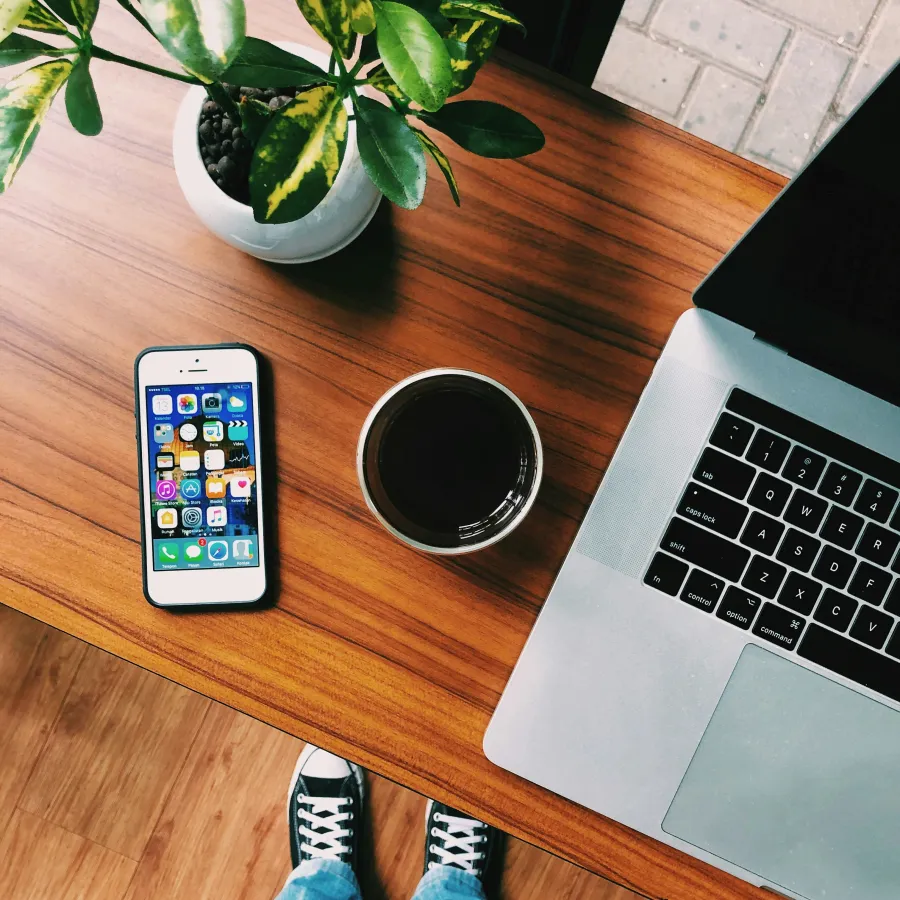 Top view of a wooden desk with a smartphone, coffee cup, laptop, and a green potted plant with someone wearing jeans and sneakers.