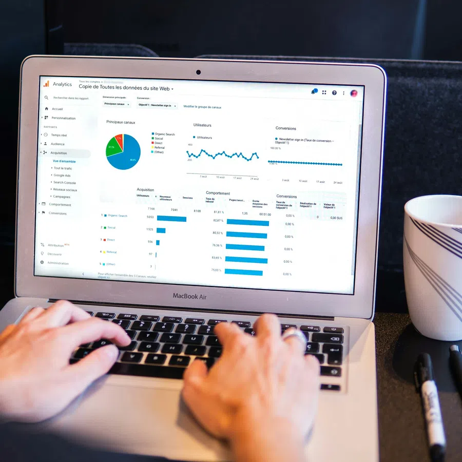 Person using a MacBook Air showing web analytics dashboard with charts, next to a striped coffee mug and pens.