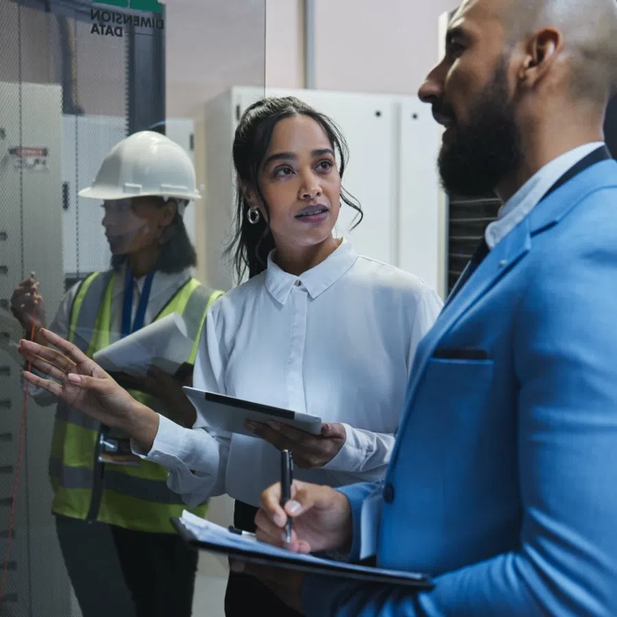 Two professionals discussing data on a screen with a worker in safety gear in the background inside a control room.