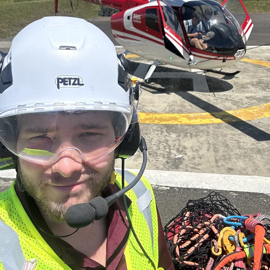 A worker wearing safety gear and headset stands near a red and white helicopter on a helipad under blue sky.