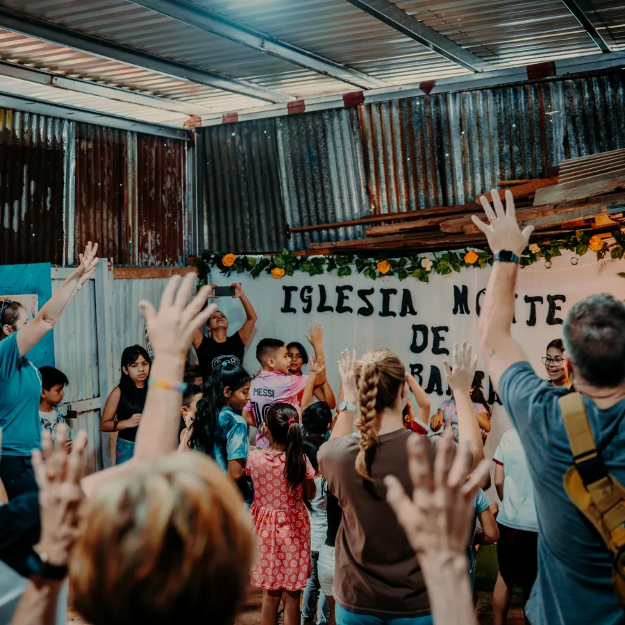 People raising hands in a lively church gathering inside a rustic metal building with a sign reading Iglesia Monte de Gracia.