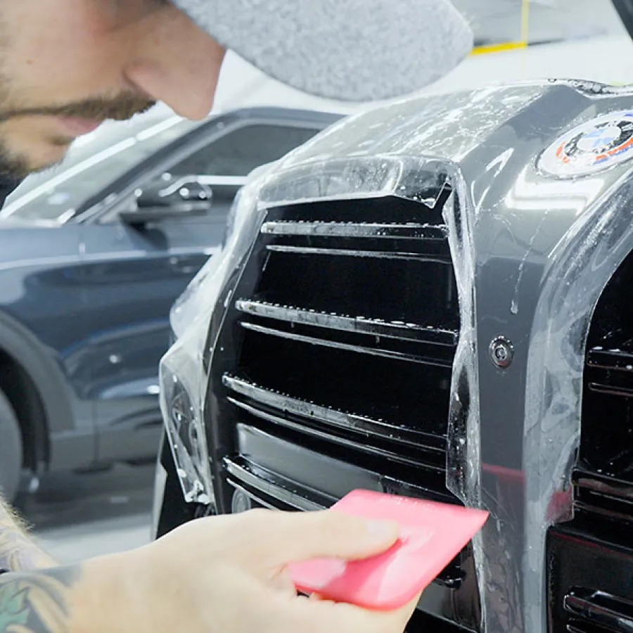 Close-up of a man applying protective film to a car's front grille using a pink squeegee in a garage.
