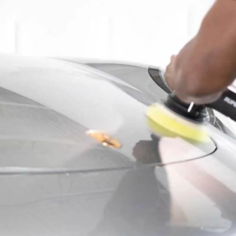 Close-up of a person polishing a silver car hood with an electric buffer in a bright garage.