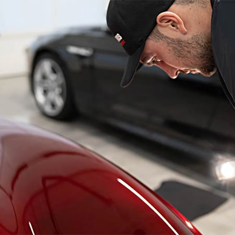 Man closely inspecting a red car's surface with a flashlight in a garage with a black car in the background