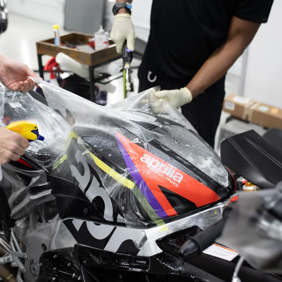 Two people applying protective film to the black Aprilia racing motorcycle with a spray bottle and tools.