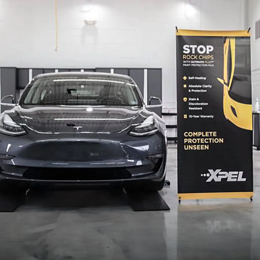 Black Tesla Model 3 parked indoors next to an XPEL banner promoting paint protection film features.