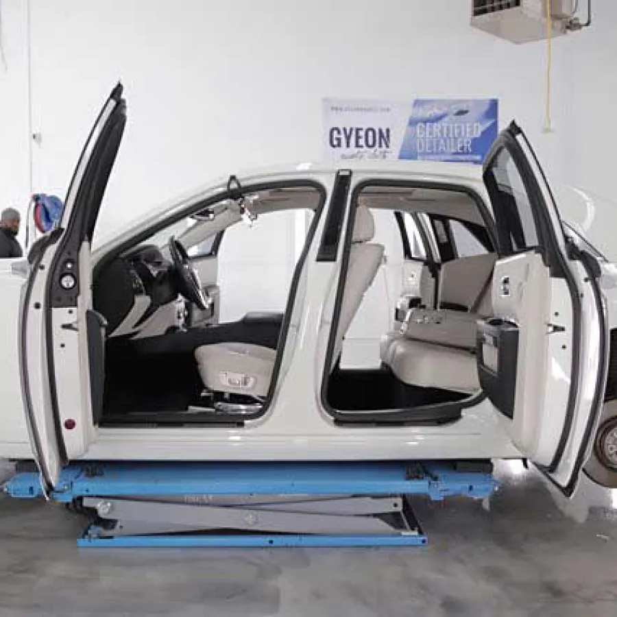 White sedan with open doors and missing wheels on a blue car lift in a garage interior.